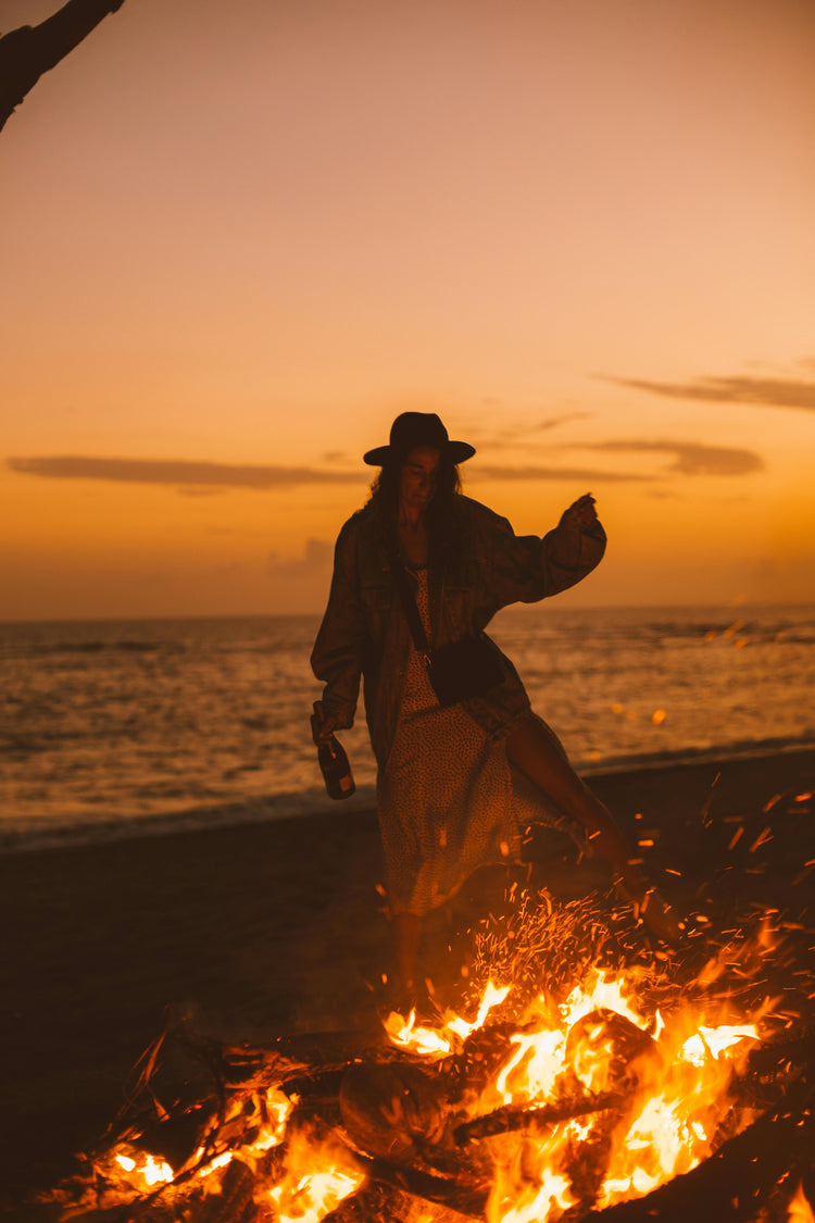 Woman behind a bonfire wearing a hat, by the beach on a beautiful sunset