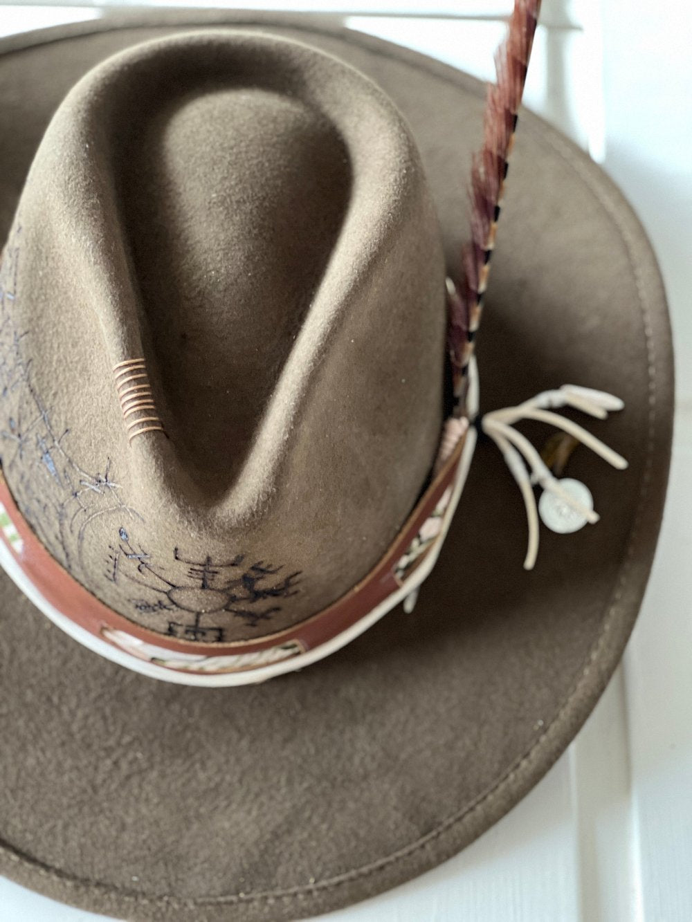 Green felt hat with design on the crown, it has a multicolor feather on the right side and a brown & white stripe at the base of the crown