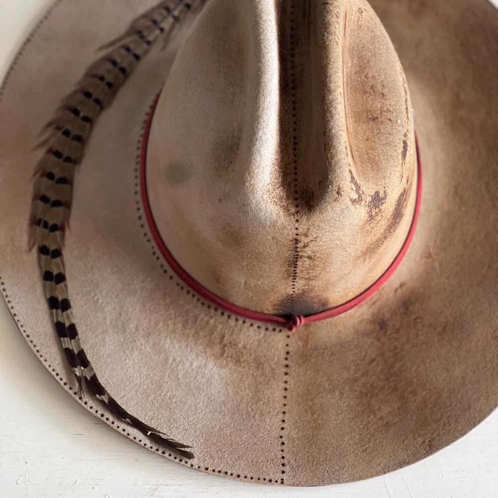 White Felt hat with dotted design markings along the left brim, a big feather on the brim spanning from top to bottom, there is a red lace.