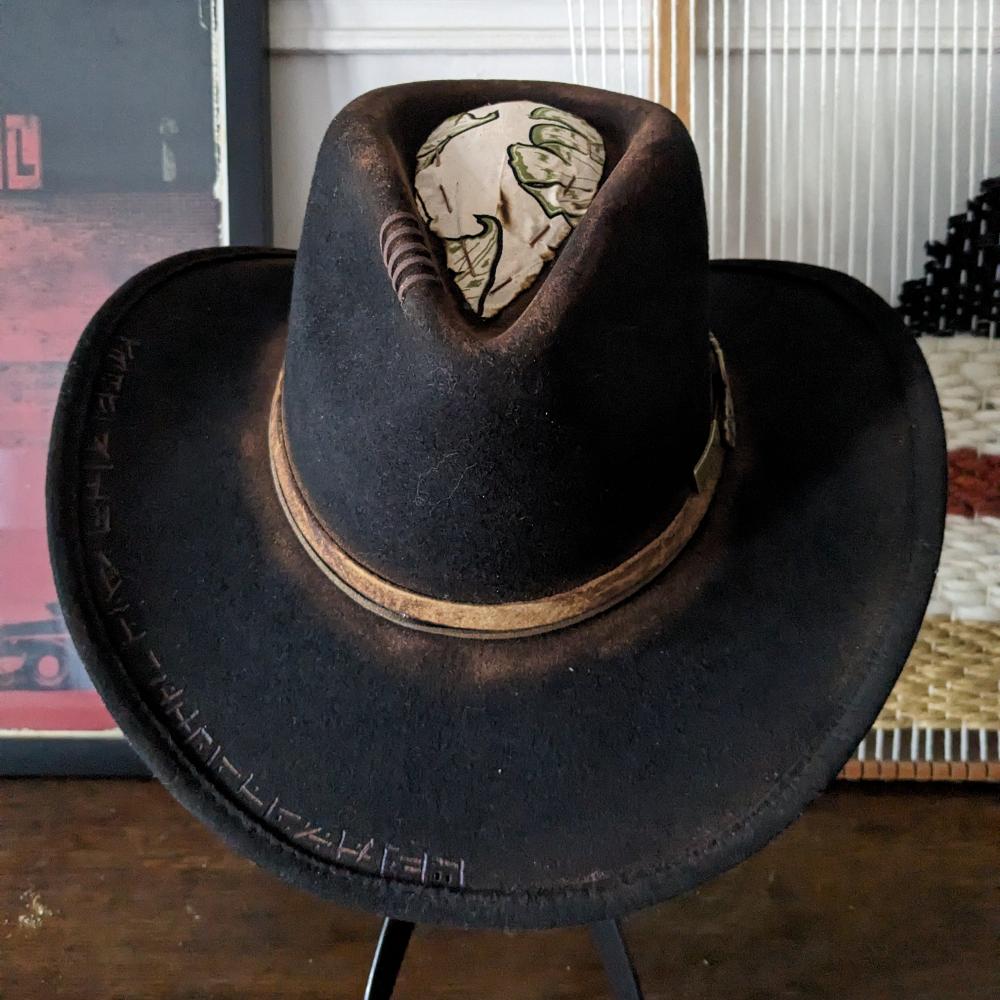 Brown felt hat on a stand, has unique design on the edge of the brim, it has a light brown stripe at the base of  the Crown