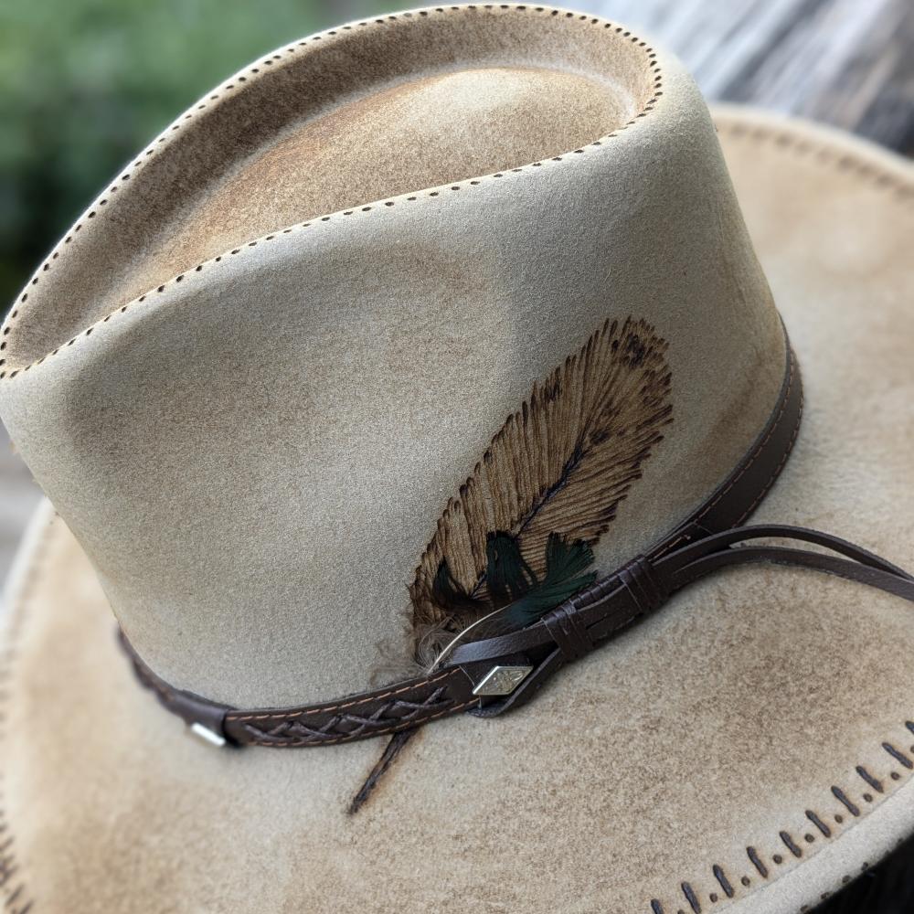Beige felt hat placed on a wooden bench; there is a leather lace at the base of the crown and markings making the hat look ruged