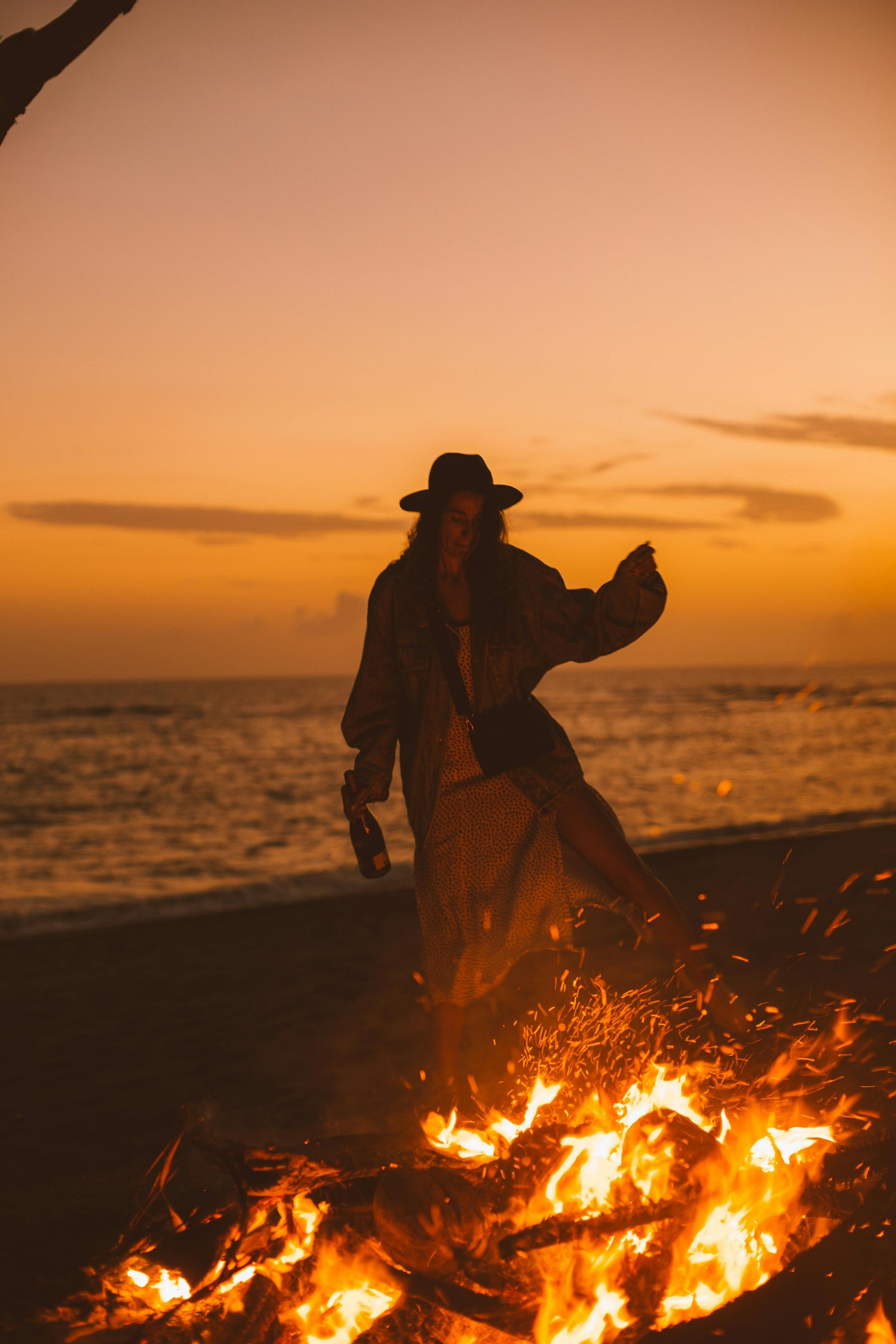 Woman behind a bonfire wearing a hat, by the beach on a beautiful sunset